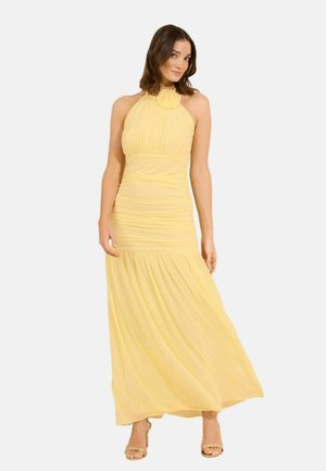 Kvinde iført en gul maxikjole med skulderstropper og plisserede detaljer samt åbne hæle, stående foran en ensfarvet hvid baggrund.