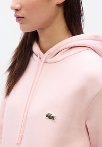 Ροζ ανοιχτόχρωμο φούτερ με κουκούλα από απαλό ύφασμα και τσέπη καγκουρό. Διαθέτει μικρό, κεντημένο πράσινο λογότυπο κροκόδειλου στο στήθος.