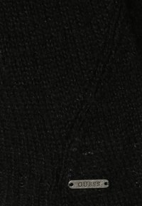Tissu tricoté noir au aspect texturé, avec une petite étiquette métallique imprimée avec "GUESS" en bas.
