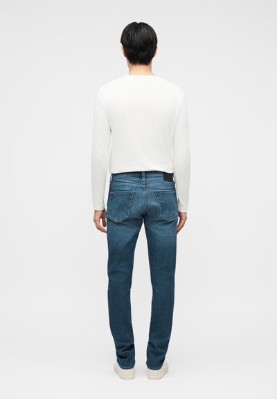 Άνδρας που βλέπει προς τα πίσω, φοράει λευκό μακρυμάνικο πουκάμισο, μπλε τζιν με πίσω τσέπες και λευκά αθλητικά παπούτσια, στέκεται σε απλό φόντο.