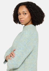 Jasnoniebieski sweter o teksturowanym wzorze, z wysokim kołnierzem i długimi rękawami, wykonany z miękkiego dzianego materiału. Model stoi bokiem, złożone ramiona.