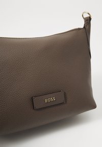 Borsa in pelle marrone con una superficie texturizzata, dotata di una placca con logo sottile in oro. Include una cerniera e un anello per attacco.