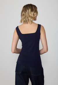 Top fără mâneci, de culoare bleumarin, cu decolteu pătrat și bretele late. Fabricat dintr-un material neted și elastic, se potrivește corpului. Se asortează cu blugi închis la culoare.