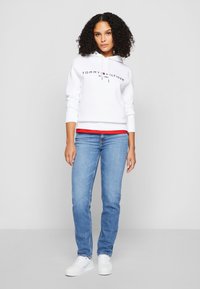 Biely mikina s kapucňou a logom "Tommy Hilfiger", modré džínsy a biele tenisky. Obsahuje kapucňu so šnúrkami a červený akcent na spodnej časti mikiny.