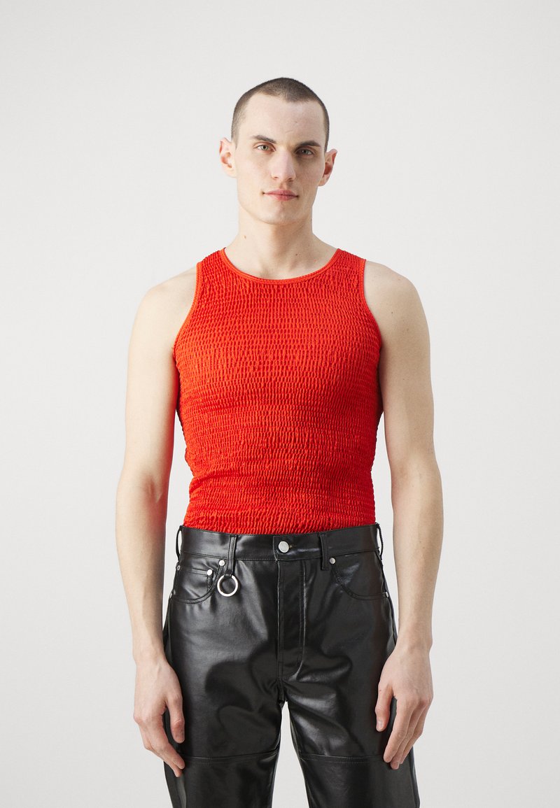 Rød teksturert tank topp med en tettsittende design, sammen med svarte skinnbukser med ringdetalj. Modellen står med fronten mot kameraet.