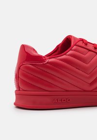 Rote Sneaker mit einer glatten, glänzenden Textur und einem Zickzackmuster im Obermaterial. Verfügt über einen gepolsterten Kragen und eine markierte Außensohle.