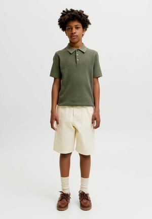 Niño con pelo rizado que lleva un polo de punto verde de manga corta, pantalones cortos beige hasta la rodilla, calcetines crema y zapatos de cuero marrón, de pie mirando hacia adelante.