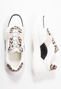 Zapatillas blancas con acentos negros y detalles de ante beige, con una sección de estampado de leopardo en el talón y suela de goma texturizada.