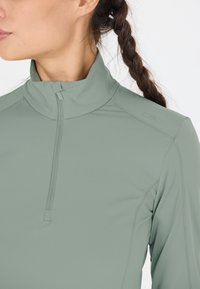 Lys grøn langærmet top med høj krave, der har en kvart lynlås og diskret logo-detalje på skulderen. Blødt, strækbart stof.