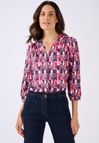 Blusa de manga larga en un patrón geométrico azul, rosa y crema; presenta cierres de botones y detalles fruncidos en los hombros. Combinada con jeans oscuros.