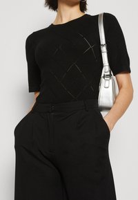 Kvinna iförd svart kortärmad topp med diamantrand, högmidjade svarta byxor och bärande en liten silverfärgad axelväska.