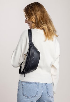 Bandolera de cuero negro con forma curva, compartimento con cremallera, correa plana y textura suave, llevada sobre un suéter blanco.