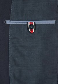 Dunkelblaues Gewebe mit strukturiertem Finish, versehen mit einer horizontalen Tasche mit einem rot-weißen gestreiften Akzent und einem schwarzen Knopfdetail.