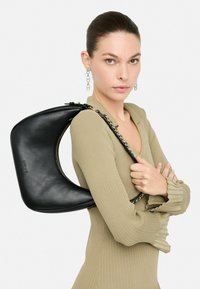 Musthave must värvitud nahast käekott, millel on kaarjas disain ja kuldsed detailid, kantud üle õla. Mudel kannab ribakujulist oliivivärvi kampsunit.