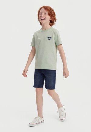 Młody chłopiec o rudych włosach uśmiecha się, idzie, nosząc jasną zieloną koszulkę z motywem palmy, ciemne dżinsowe szorty i białe trampki.