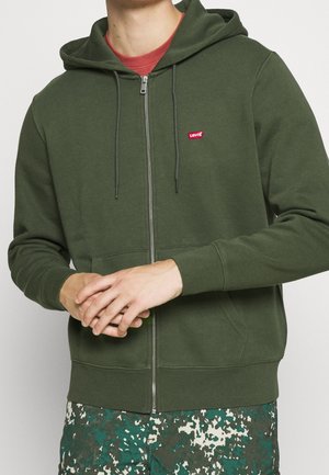 Mężczyzna w zielonej bluzie Levi's na zamek, na czerwonej koszulce i zielonych, białych i niebieskich szortach w wzory, z rękami splecionymi z przodu.