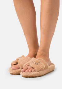 Fuzzy beige slides with a soft texture, featuring a strap adorned with a square metallic accent and a rubber sole.