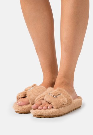 Fuzzy beige slides with a soft texture, featuring a strap adorned with a square metallic accent and a rubber sole.