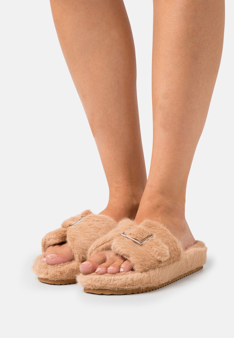 Fuzzy beige slides with a soft texture, featuring a strap adorned with a square metallic accent and a rubber sole.