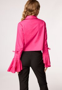 Camicetta fucsia a maniche lunghe con polsini svasati e dettagli a fiocco, abbinata a pantaloni neri attillati. Texture liscia.