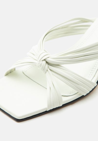 Λευκό σανδάλι με τετράγωνη μύτη και πολλαπλούς λεπτούς ιμάντες δεμένους σε διακοσμητικό κόμπο μπροστά.