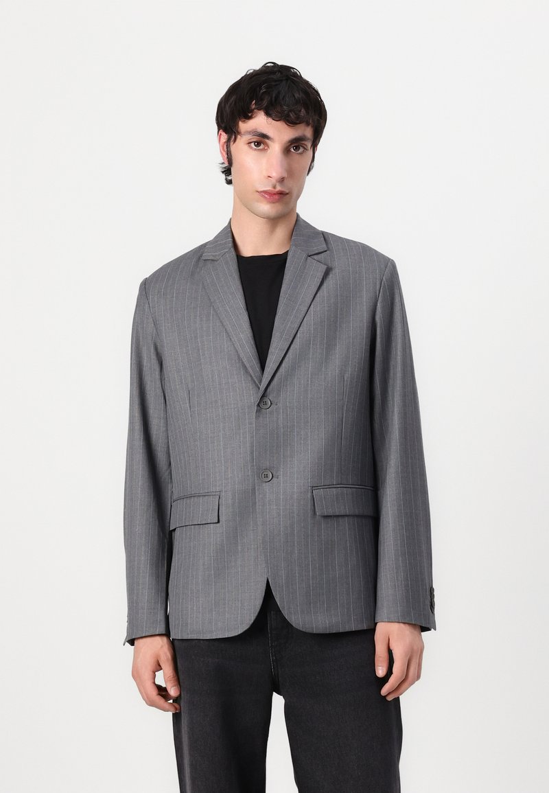 Gray striped blazer with a two-button closure, notched lapels, and two front pockets. Smooth texture, relaxed fit, paired with a black shirt.