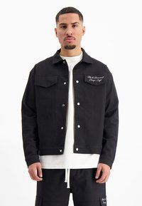 Zwarte denimjack met een kraag, drukknopen, dubbele voorzakken en een witte geborduurde logo op de borst.