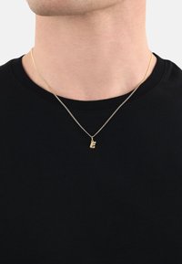 Collana d'oro con un piccolo pendente a forma di "E" stilizzata, appesa a una delicate catena. Indossata su una T-shirt nera a costine.