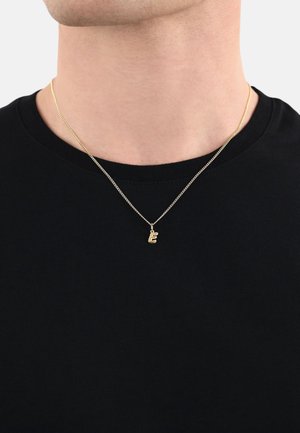 Gouden ketting met een kleine, gestileerde "E" hanger, hangend aan een delicate ketting. Draagbaar over een zwart, geribbeld T-shirt.