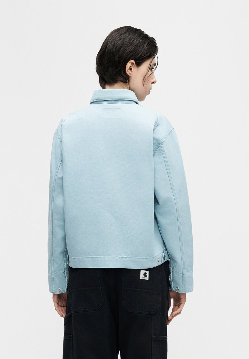 Personne aux cheveux courts et foncés, vue de dos, portant une veste bleu clair et un pantalon noir avec un patch de marque visible sur la poche.