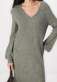 Robe pull verte à encolure en V en matériau tricot doux, dotée de manches longues et d'une coupe décontractée, avec une finition côtelée et une texture subtile.