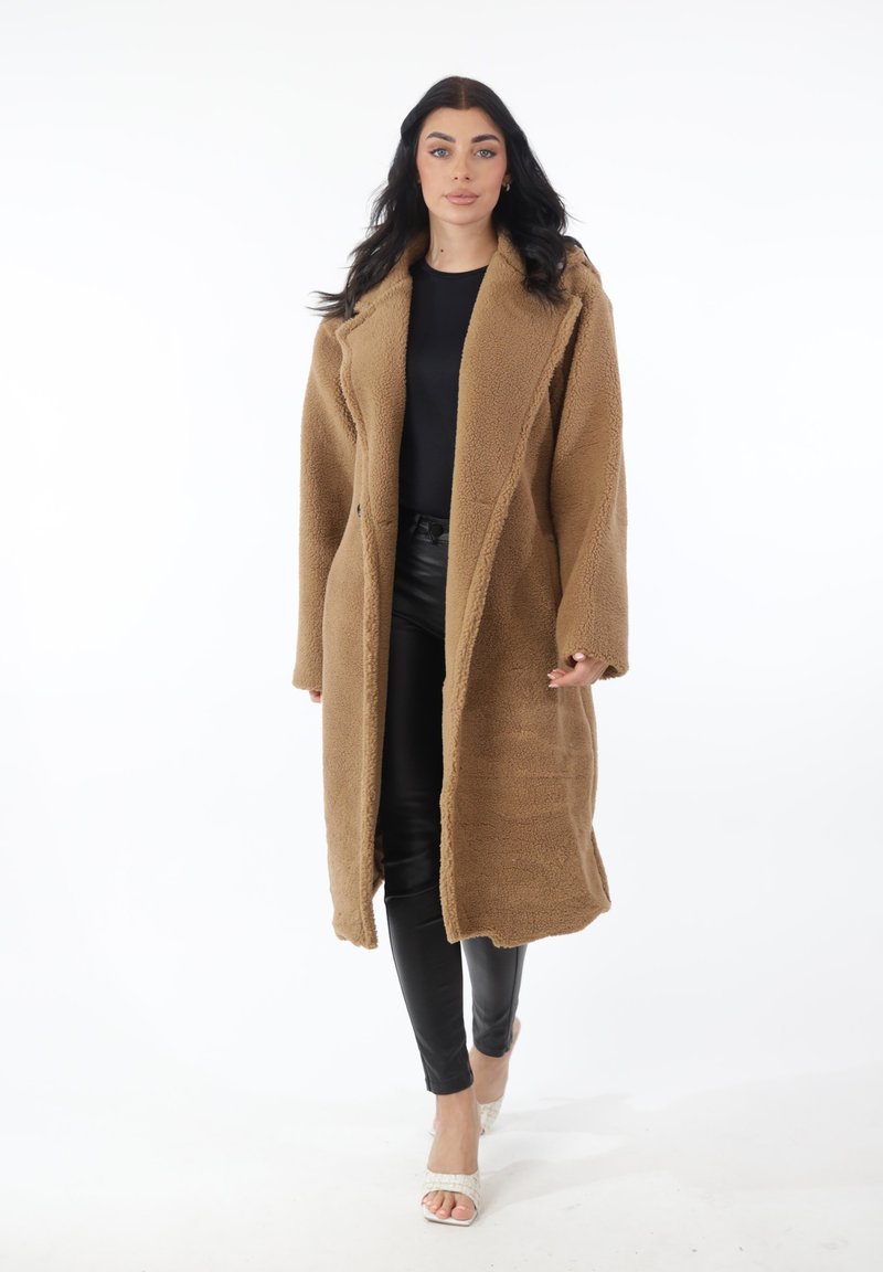 Mujer caminando hacia adelante con un abrigo largo de peluche marrón, camiseta negra, pantalones de cuero negros y tacones blancos abiertos, sobre un fondo blanco.