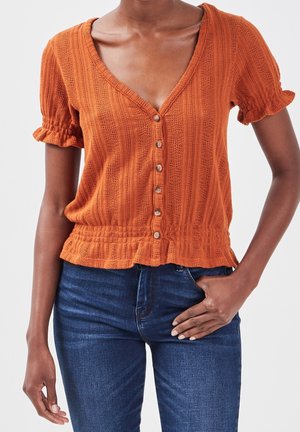 Femme portant un chemisier orange texturé à manches courtes boutonné avec des poignets à volants et un jean bleu foncé, main posée sur la hanche.