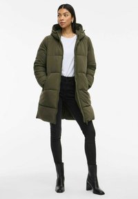 Giacca puffer verde oliva con cappuccio, dotata di chiusura zip, design trapuntato e due tasche laterali. Indossata sopra una camicia bianca e jeans neri.