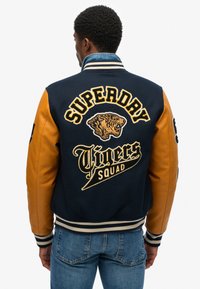 Veste varsity en tissu bleu marine avec des manches en cuir orange, logo de tigre brodé et texte "SUPERDRY TIGERS SQUAD" dans le dos.