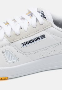 Sneakers bianchi realizzati in pelle liscia con un accento in suede, presentano dettagli perforati, lacci bianchi e una suola gialla. Presente il logo Reebok.