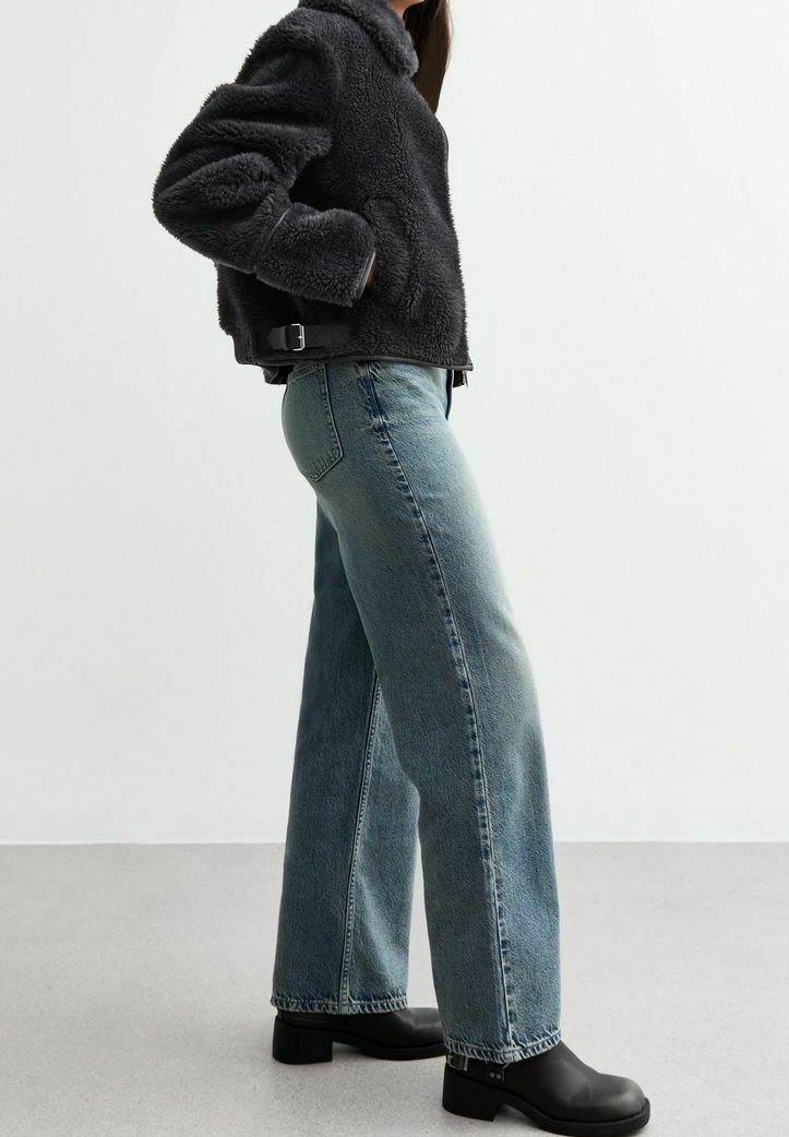 Femme portant une veste noire duveteuse, un jean droit bleu clair et des bottines noires, debout devant un fond clair.