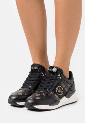 Black and brown patterned Guess sneakers with white wedge soles worn on bare legs against a white background.