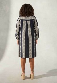 Löysä musta ja kerma valkoinen mekko, jossa on geometrinen kuvio, pitkät hihat ja puolipitkä helma. Varoitsee teksturoituja yksityiskohtia hihojen varrella.