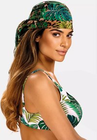 Strój kąpielowy z zielonymi liściami na białym tle; modelka nosi pasującą zielono-brązową chustkę na głowie, widoczne falujące włosy.