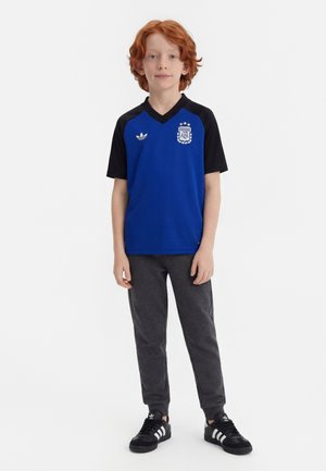 Ragazzo dai capelli rossi in piedi che indossa una maglia da calcio dell'Argentina blu e nera, pantaloni grigio scuro e scarpe da ginnastica Adidas nere con strisce bianche.