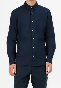 Camicia a maniche lunghe blu navy con colletto abbottonato, taschino frontale e bottoni bianchi. Il tessuto sembra liscio e leggermente strutturato.