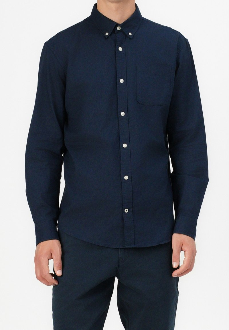 Camicia a maniche lunghe blu navy con colletto abbottonato, taschino frontale e bottoni bianchi. Il tessuto sembra liscio e leggermente strutturato.