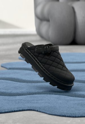 Melns aplikts slip-on apavs ar sprādzes siksniņu, bieza, robusta zole, novietots uz zilas teksturētas virsmas ar izplūdušu mīkstinātu fonu.