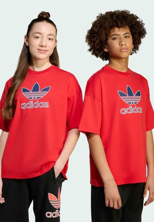 Due adolescenti indossano magliette rosse Adidas e pantaloni neri Adidas, in piedi uno accanto all'altro davanti a uno sfondo uniforme.