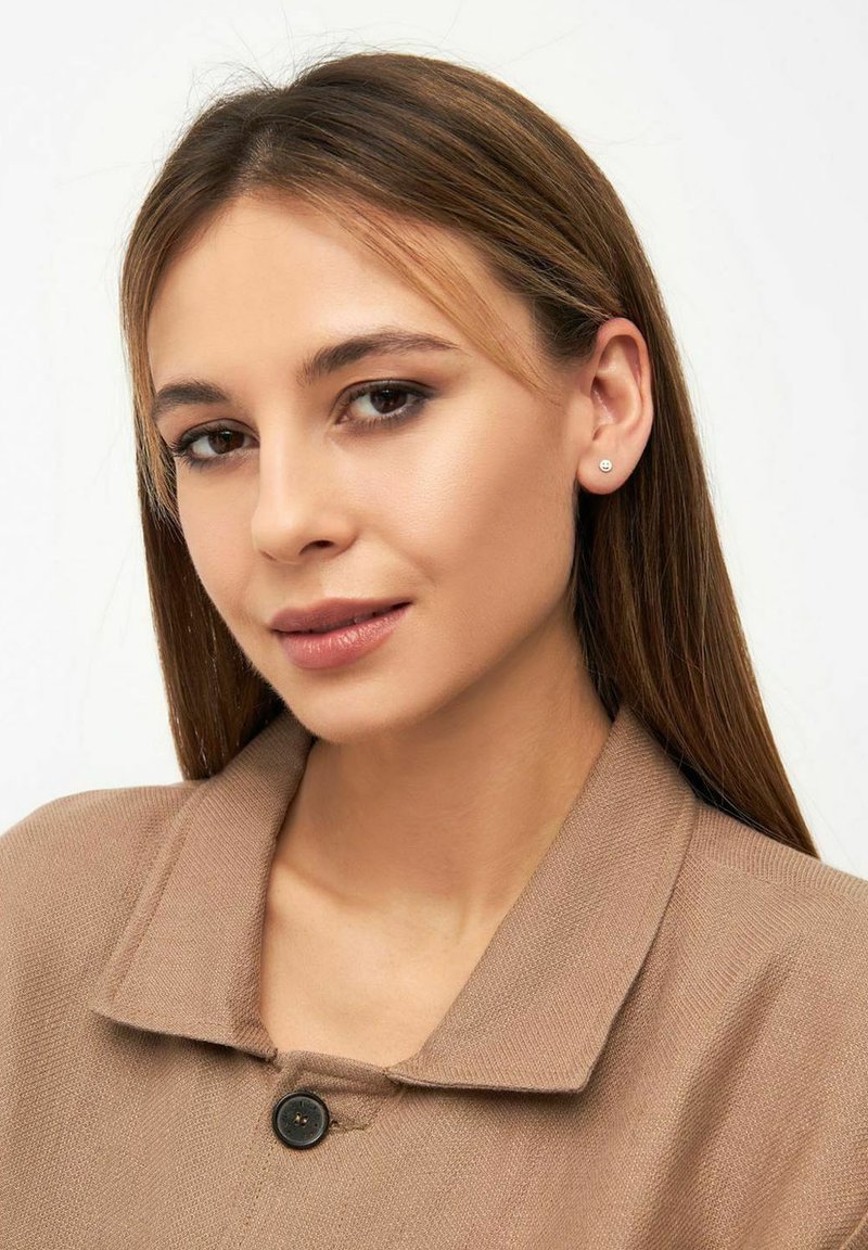 Kremkrāsas jaciņa ar pogām un apkakli, ko valkā sieviete ar taisniem, brūniem matiem. Vienkāršas auskaru püšas un minimāla grima.