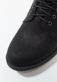 Gros plan sur une chaussure noire en cuir à lacets avec une surface texturée et des œillets métalliques, sur fond blanc.