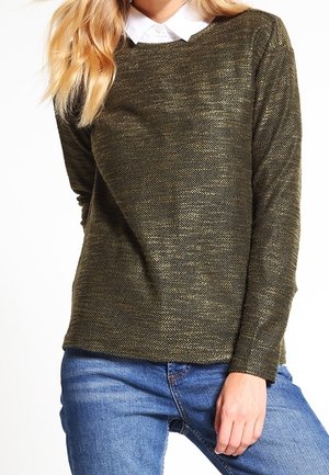 Kvinde iført en olivengrøn striktrøje over en hvid skjorte med krave og blå jeans, poserer med den ene hånd hvilende på låret.