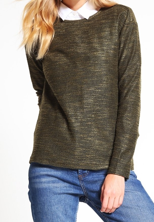 Femme portant un pull en maille vert olive par-dessus une chemise blanche à col, avec un jean bleu, posant avec une main reposant sur sa cuisse.