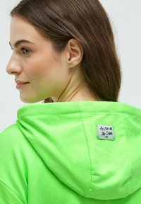 Leuchtend grüner Kapuzenpullover mit weicher Textur, ausgestattet mit einer Kapuze und einem aufgenähten Etikett auf der Rückseite, auf dem "So frei wie der Ozean" steht.